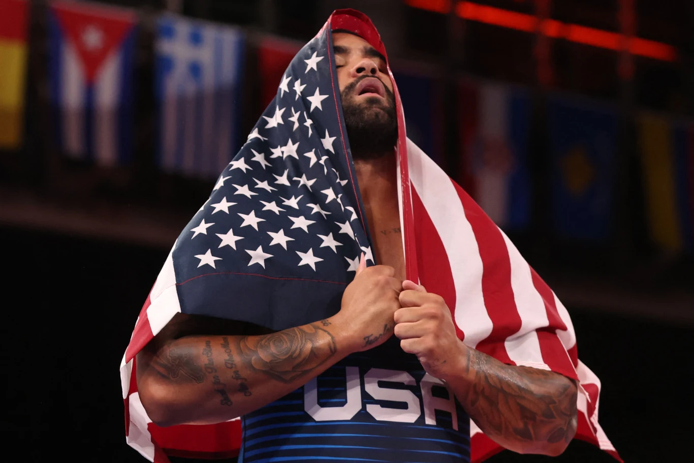 Gable Steveson Zawodnik ubrany w strój sportowy z napisem USA trzyma na sobie flagę Stanów Zjednoczonych, która okrywa mu głowę i ramiona, w tle widać inne flagi państwowe.