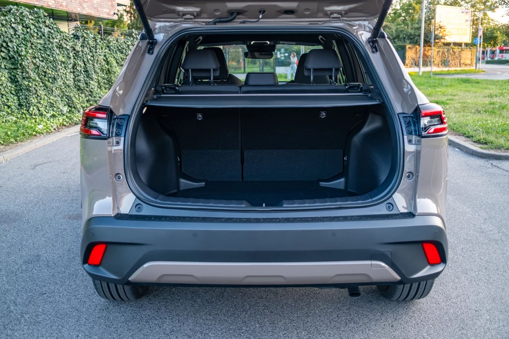 473 l pojemności i regularne kształty. Bagażnik to zaleta Toyoty Corolli Cross Otwarty bagażnik nowoczesnego samochodu typu SUV stojącego na ulicy, wnętrze bagażnika jest puste i dobrze widoczne, w tle fragment zielonego żywopłotu oraz asfaltowa droga