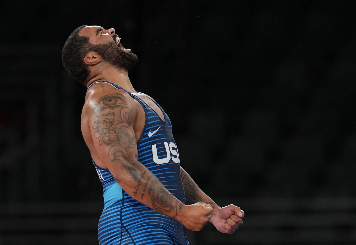 Gable Steveson Sportowiec w niebieskim stroju z widocznym napisem USA, stojący bokiem i wyrażający intensywne emocje, z zaciśniętymi pięściami i przechyloną do tyłu głową, z mocno widocznymi tatuażami na ramieniu.