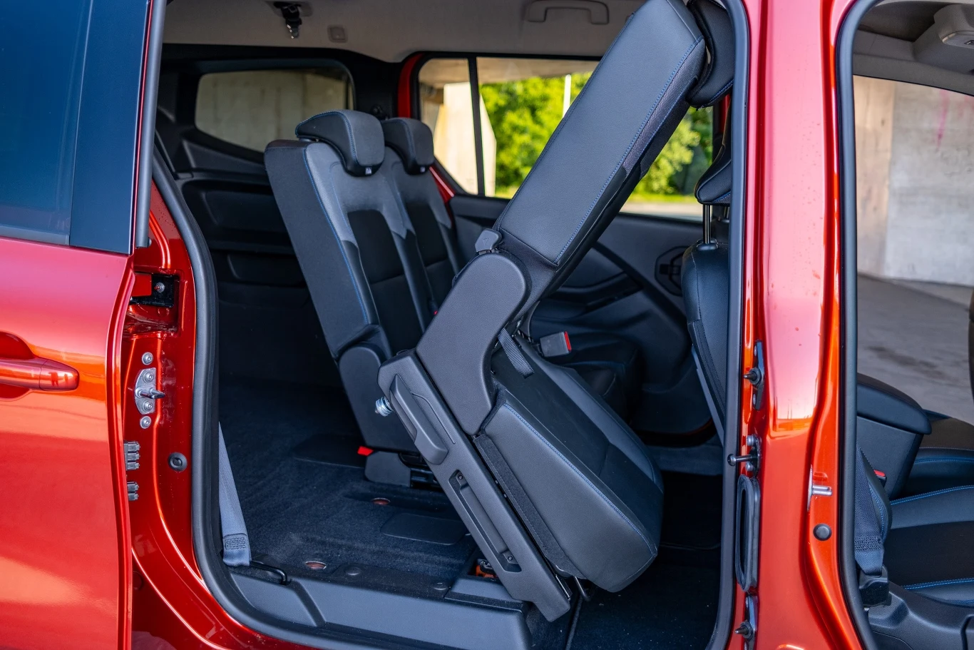 Wnętrze czerwonego samochodu z otwartymi drzwiami przesuwanymi, środkowy rząd foteli podniesiony w celu ułatwienia dostępu do tylnej części pojazdu, wyraźnie widoczna przestrzeń na nogi i tapicerka.