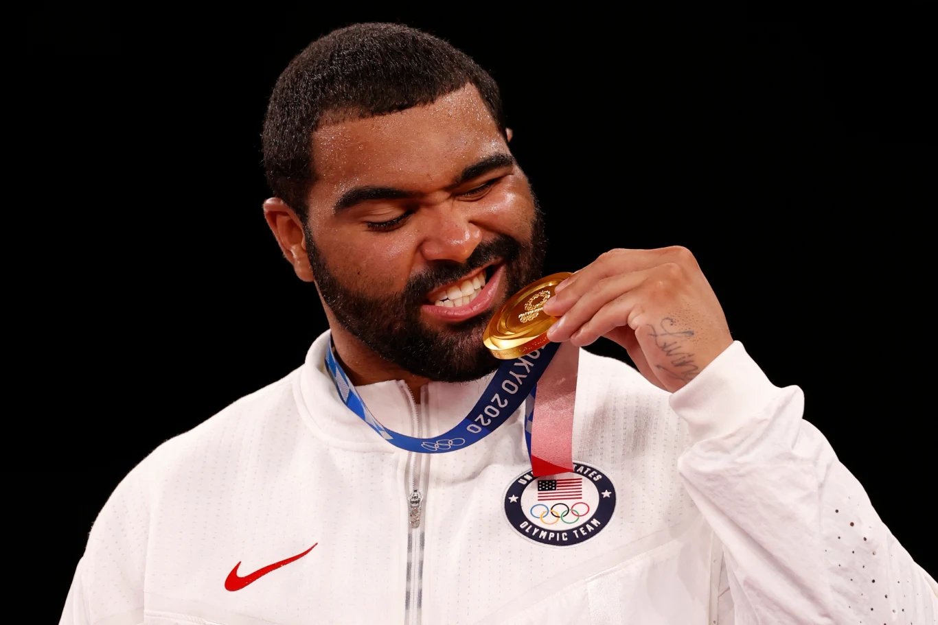 Gable Steveson Sportowiec ubrany w biały dres z logo reprezentacji USA gryzie złoty medal olimpijski, a jego twarz wyraża radość i dumę. Czarny, jednolity background podkreśla główny temat zdjęcia.