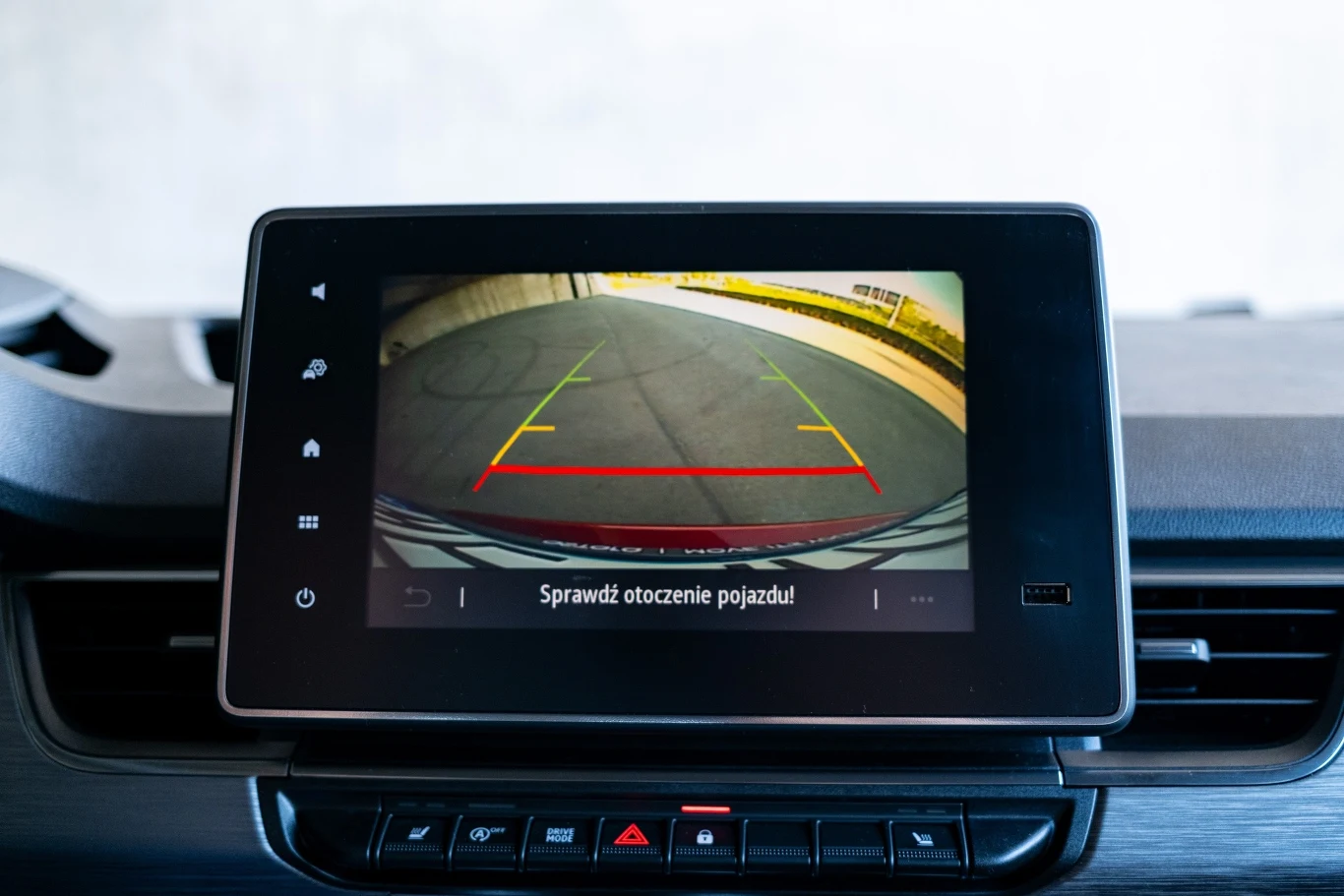 Ekran systemu cofania w samochodzie wyświetlający obraz z kamery cofania oraz kolorowe linie pomocnicze służące kierowcy do parkowania, z widocznym napisem 'Sprawdź otoczenie pojazdu'.