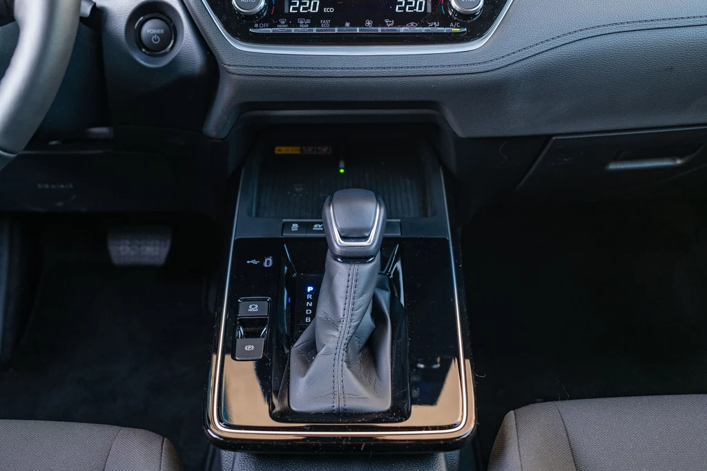 Konsola środkowa to największa nowość we wnętrzu zmodernizowanej Toyoty Corolli Cross Dźwignia automatycznej skrzyni biegów wraz z przyciskami sterującymi, otoczona czarną, błyszczącą konsolą centralną samochodu, elementy deski rozdzielczej i fragment kierownicy widoczne w górnej części kadru.