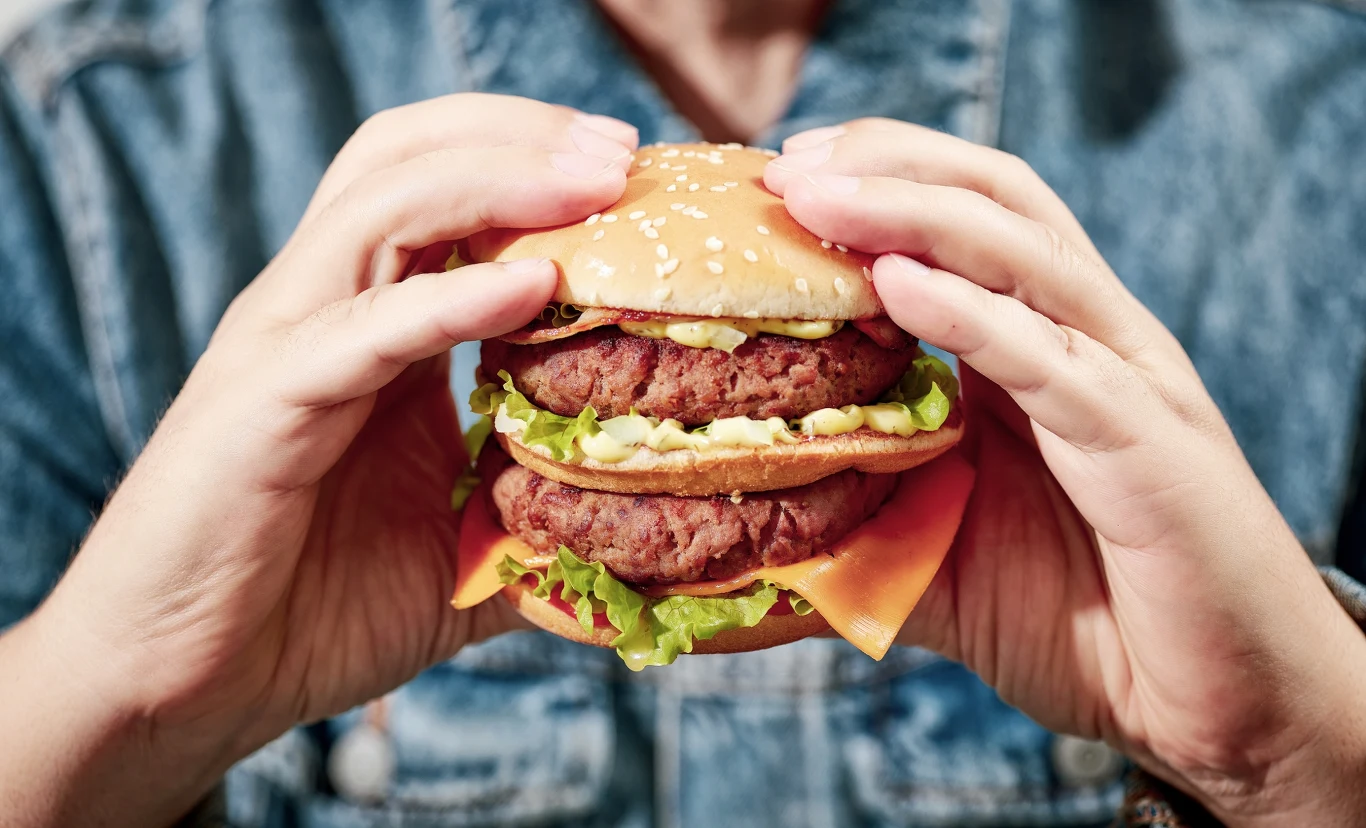 Duży hamburger z dwoma soczystymi kotletami, plasterkiem żółtego sera, liściem sałaty, oraz dodatkami, trzymany w obu dłoniach przez osobę ubraną w jeansową kurtkę.