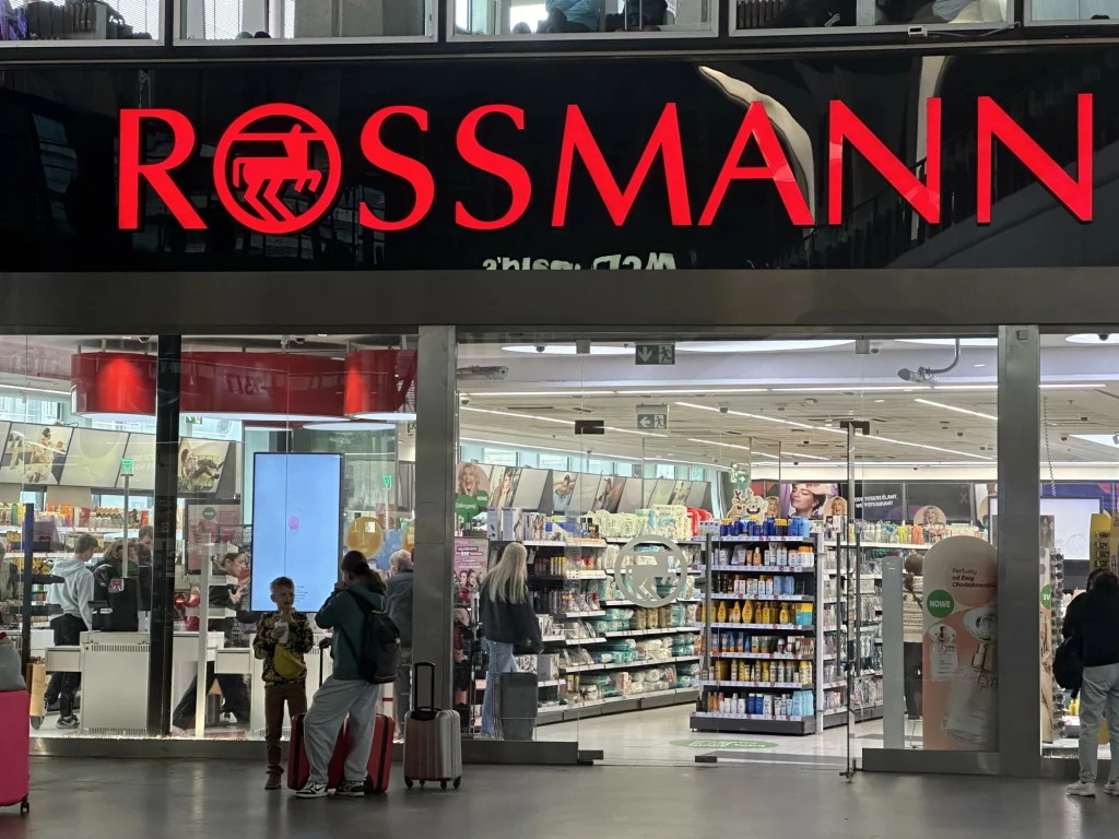 Wejście do sklepu Rossmann z dużym, podświetlonym szyldem oraz witryną, przez którą widoczny jest asortyment sklepu i klienci robiący zakupy, z przodu dwie postacie z walizkami rozmawiające przed wejściem.