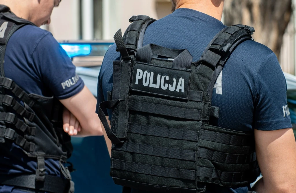 Dwóch policjantów w kamizelkach taktycznych, z widocznym napisem 'Policja', stojących obok policyjnego radiowozu.