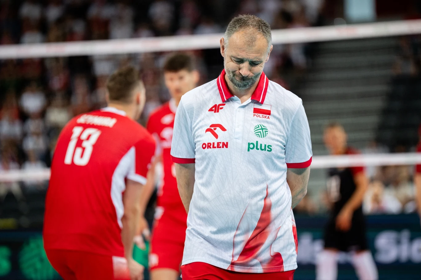 Nikola Grbić Trener w białej koszulce z logotypami sponsorów stoi ze spuszczoną głową, w tle siatkarze w czerwonych strojach, rozmyte trybuny pełne kibiców.