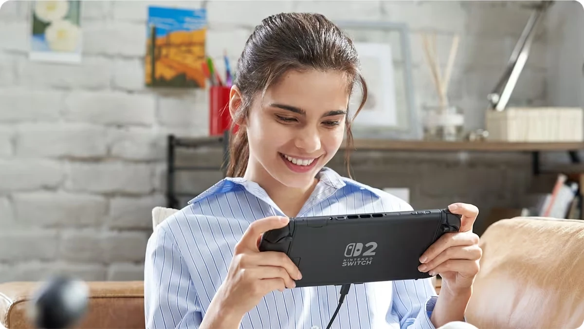 Nintendo Switch 2 Uśmiechnięta młoda kobieta w piżamie siedzi na kanapie w nowocześnie urządzonym pokoju i gra na konsoli Nintendo Switch, trzymając ją obiema rękami.