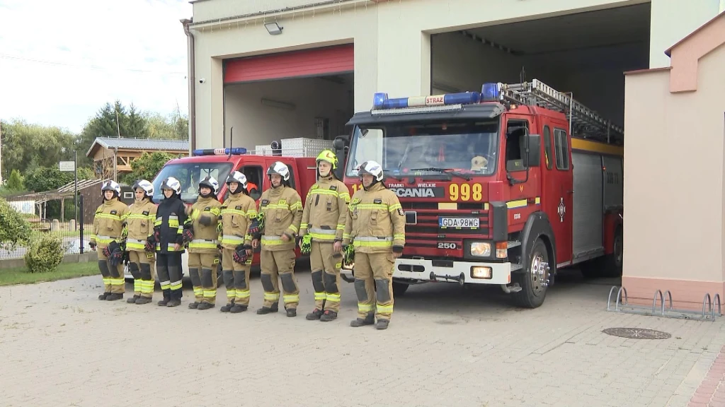 Strażacy oddali hołd zmarłemu druhowi Grupa strażaków w pełnym umundurowaniu stojąca w szeregu przed remizą strażacką, obok dwóch wozów strażackich zaparkowanych przy otwartych bramach garażu.