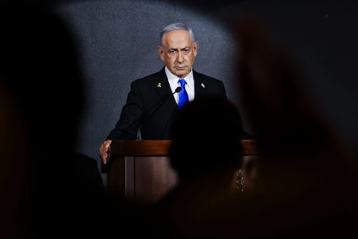 Binjamin Netanyahu deklaruje gotowość do negocjacji Mężczyzna w garniturze stojący za mównicą podczas oficjalnego wystąpienia, oświetlony reflektorem na tle ciemnej ściany, pierwszy plan nieostry, postać skupiona, z poważnym wyrazem twarzy.