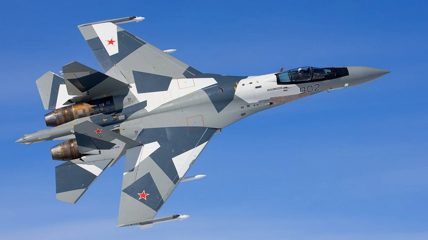 Duża dostawa najnowszych myśliwców Su-35S w Rosji Myśliwiec wojskowy w nowoczesnym, szaro-niebieskim kamuflażu z widocznymi czerwonymi gwiazdami na skrzydłach i kadłubie, lecący na tle nieba.