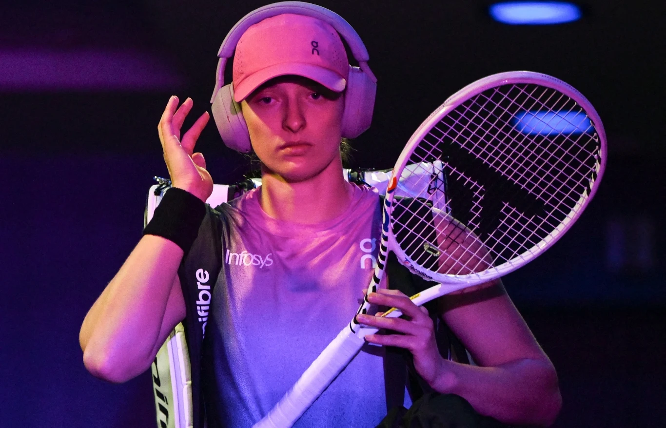 Iga Świątek Sportowiec w stroju tenisowym z rakietą na ramieniu i słuchawkami na uszach, skupiony, w trakcie przechodzenia przez oświetlone różowym światłem pomieszczenie