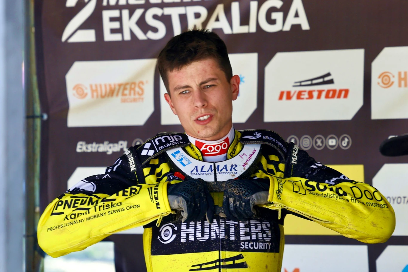 Młody żużlowiec w sportowym kombinezonie oparty o barierkę, w tle widoczne tablice z logotypami sponsorów drugiej ligi żużlowej