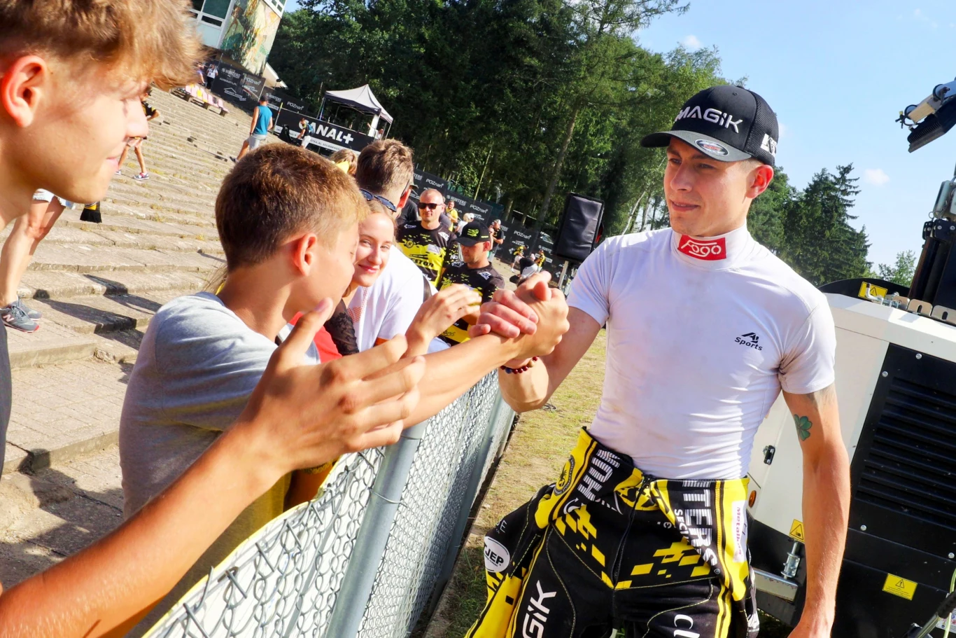 Zawodnik sportów motorowych ubrany w kombinezon wyścigowy ściska dłoń młodego kibica przy ogrodzeniu, w tle widoczni inni kibice oraz zielone drzewa.