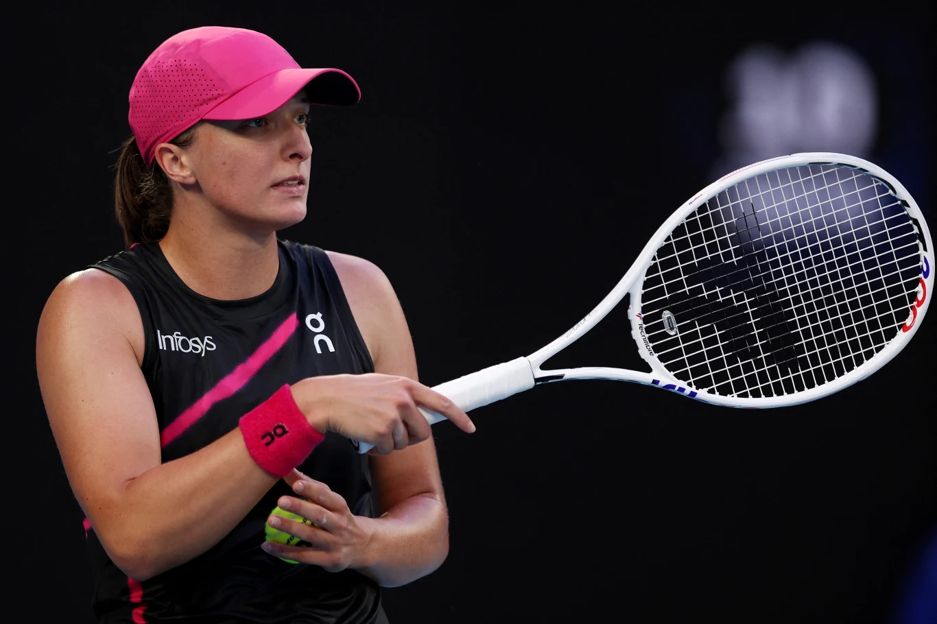 Iga Świątek Tenisistka ubrana w czarną koszulkę sportową z różowymi akcentami oraz różową czapkę trzyma rakietę tenisową i piłkę, widoczna na czarnym tle kortu.