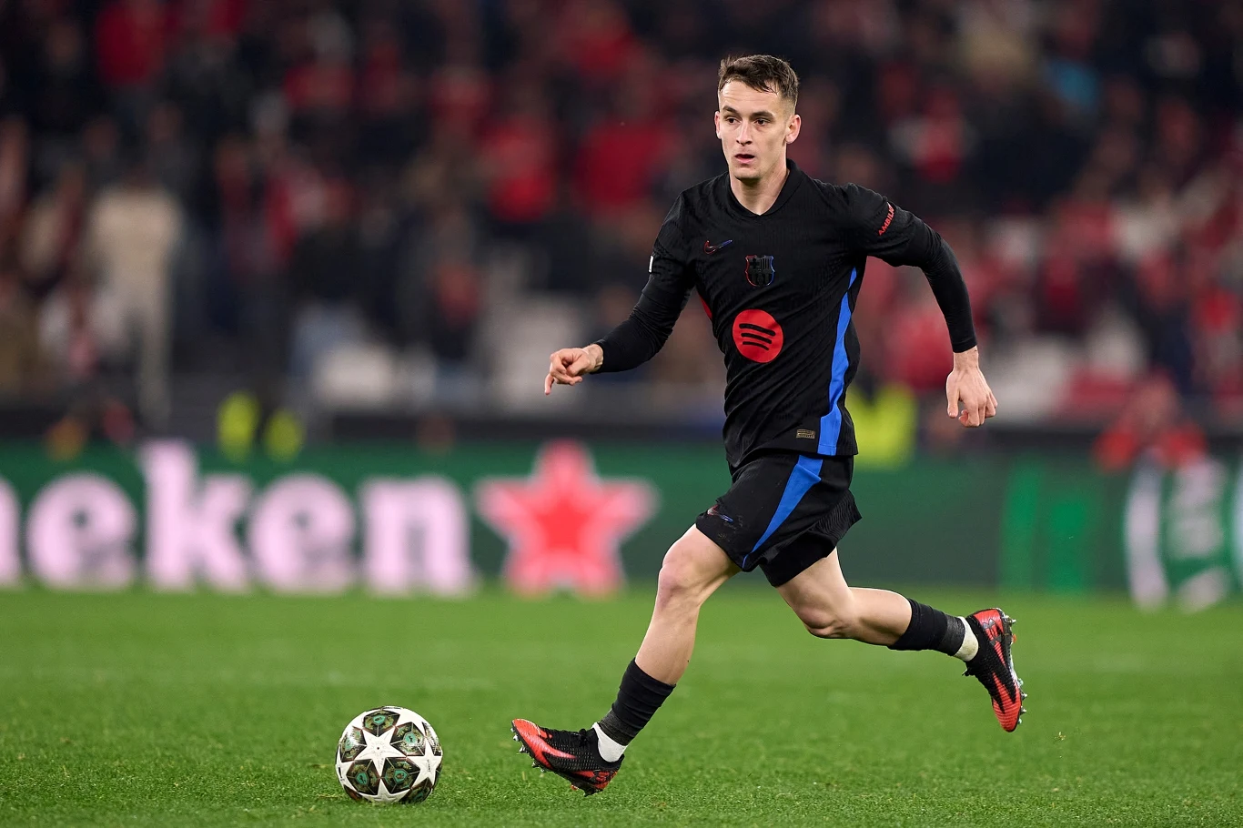 Piłkarz w czarno-niebieskim stroju biegnie z piłką na murawie stadionu, w tle widoczni kibice oraz rozmyta reklama Heineken.