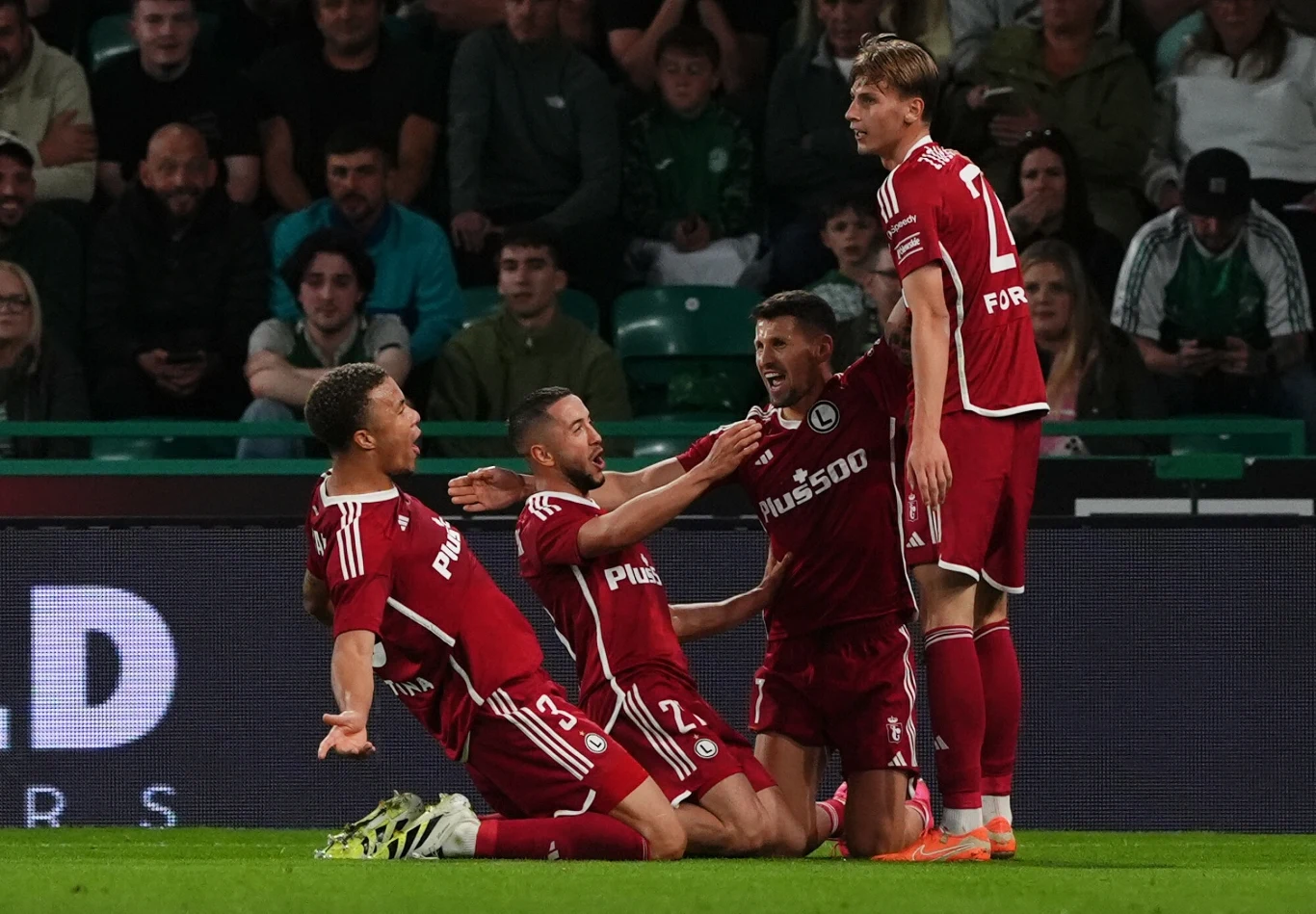 Czterech piłkarzy w czerwonych strojach celebruje zdobycie bramki na murawie stadionu, kibice obserwują ich z trybun