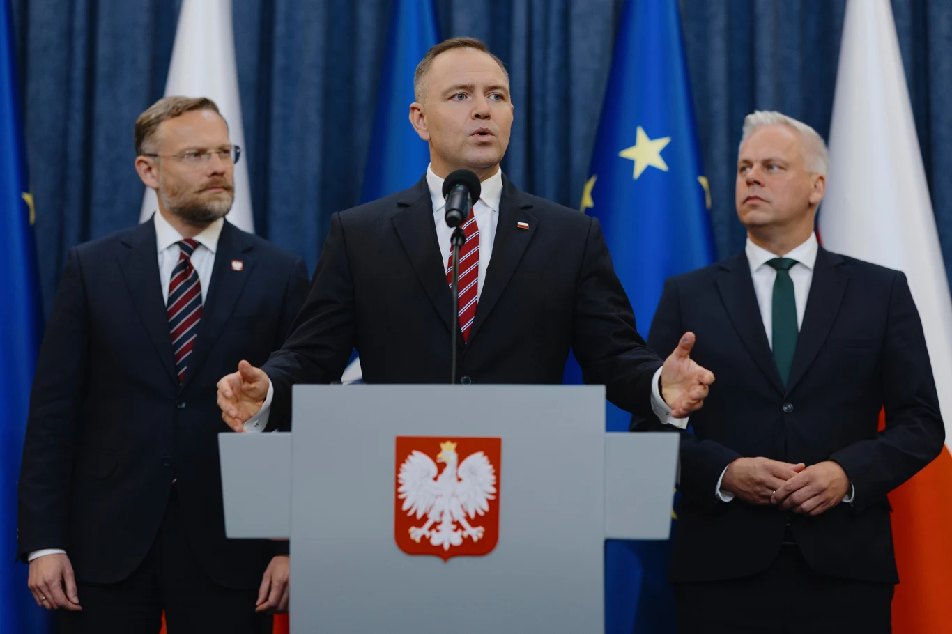 Zbigniew Bogucki, prezydent Karol Nawrocki oraz Karol Rabenda Mężczyzna wygłaszający przemówienie w otoczeniu dwóch innych mężczyzn, stojących po bokach, na tle flag Polski oraz Unii Europejskiej, przy mównicy z godłem państwowym.