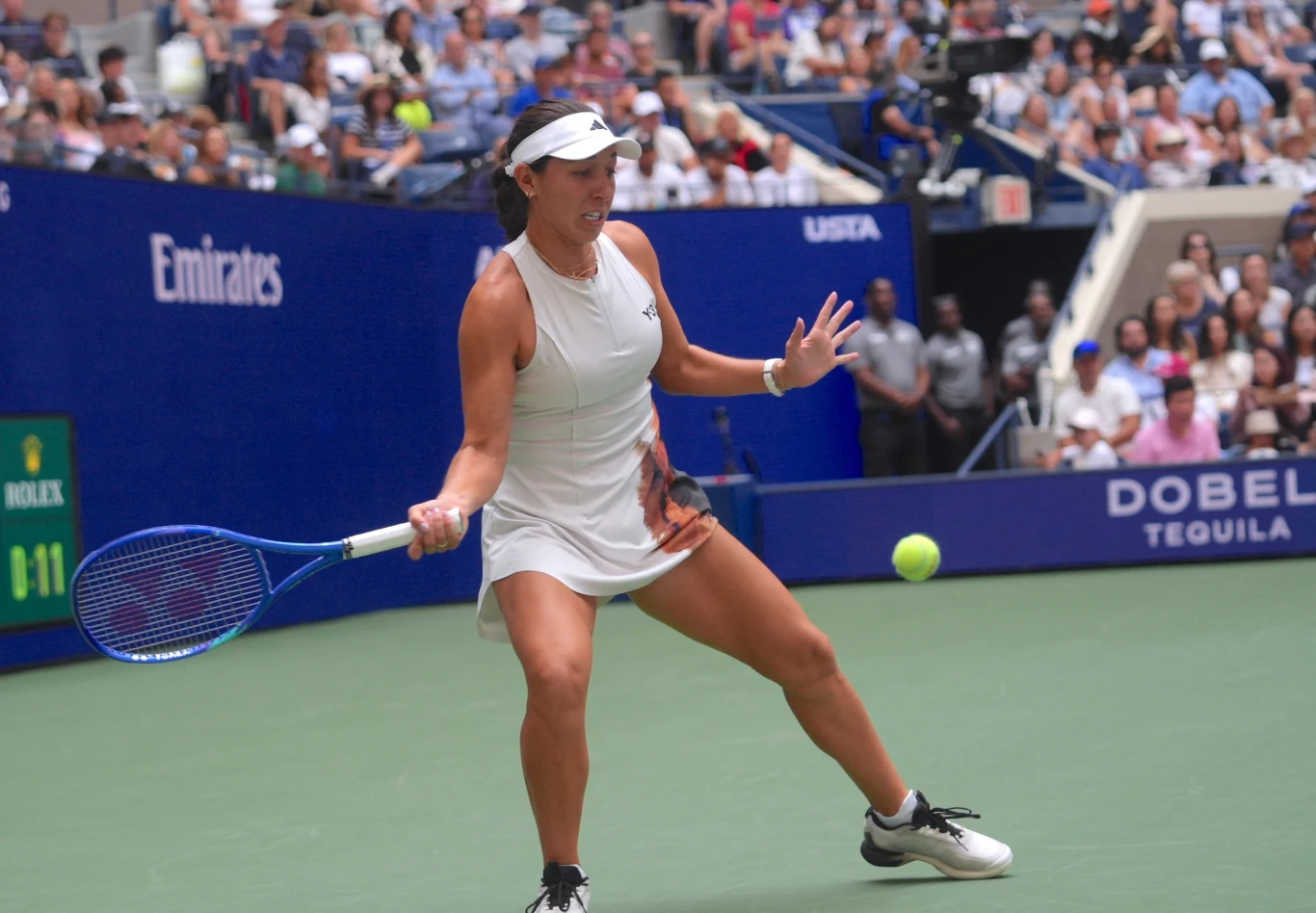 Zawodniczka tenisowa uderza piłkę podczas meczu na korcie, skoncentrowana na grze, w tle widoczna publiczność oraz elementy oznaczeń turniejowych.