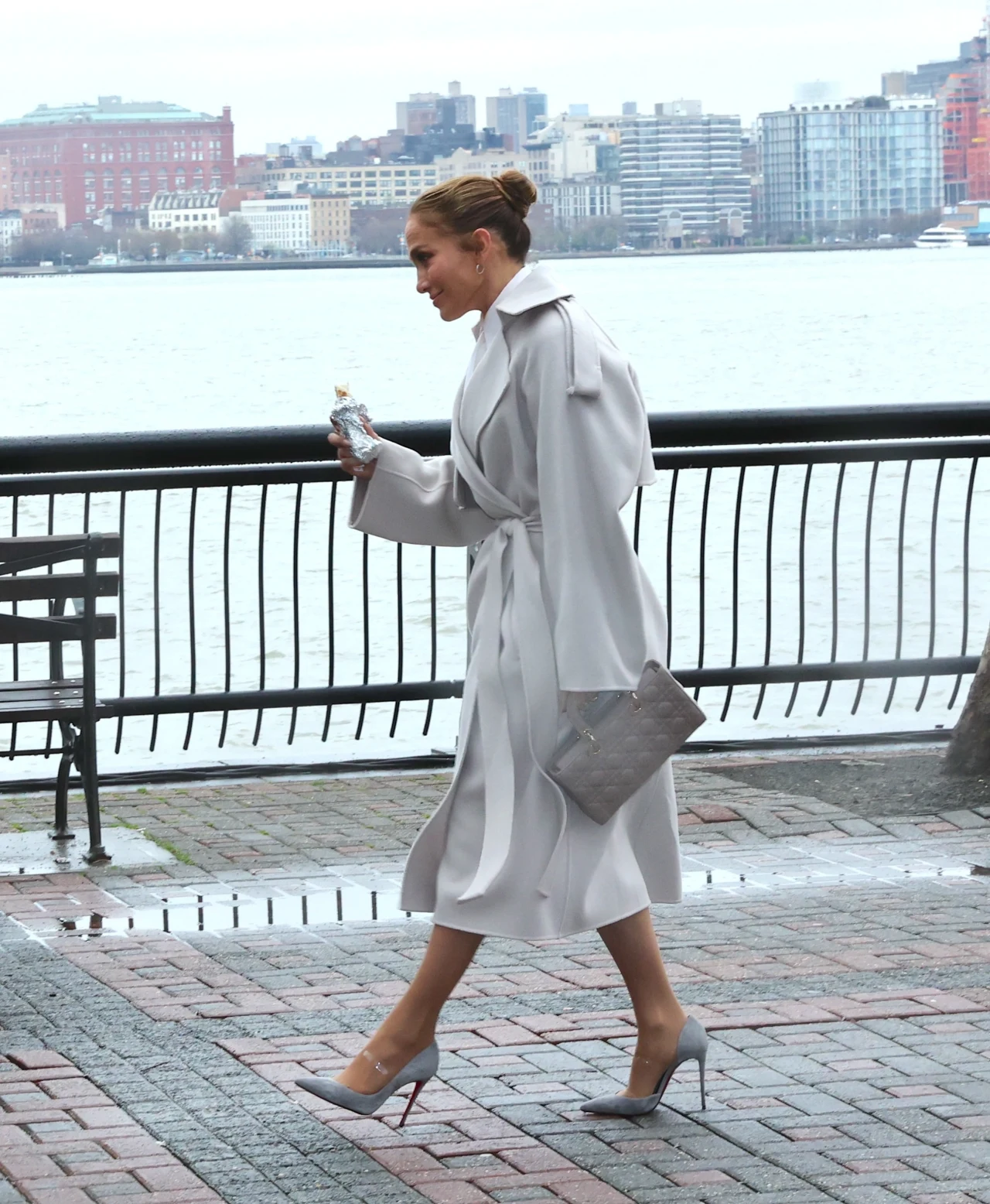 Jennifer Lopez w idealnym trenczu na jesień Elegancka kobieta ubrana w jasny płaszcz, wysokie szpilki i spinkę do włosów przechodzi chodnikiem przy nadbrzeżu, trzymając w ręku torebkę i butelkę, w tle panorama miasta po drugiej stronie rzeki.