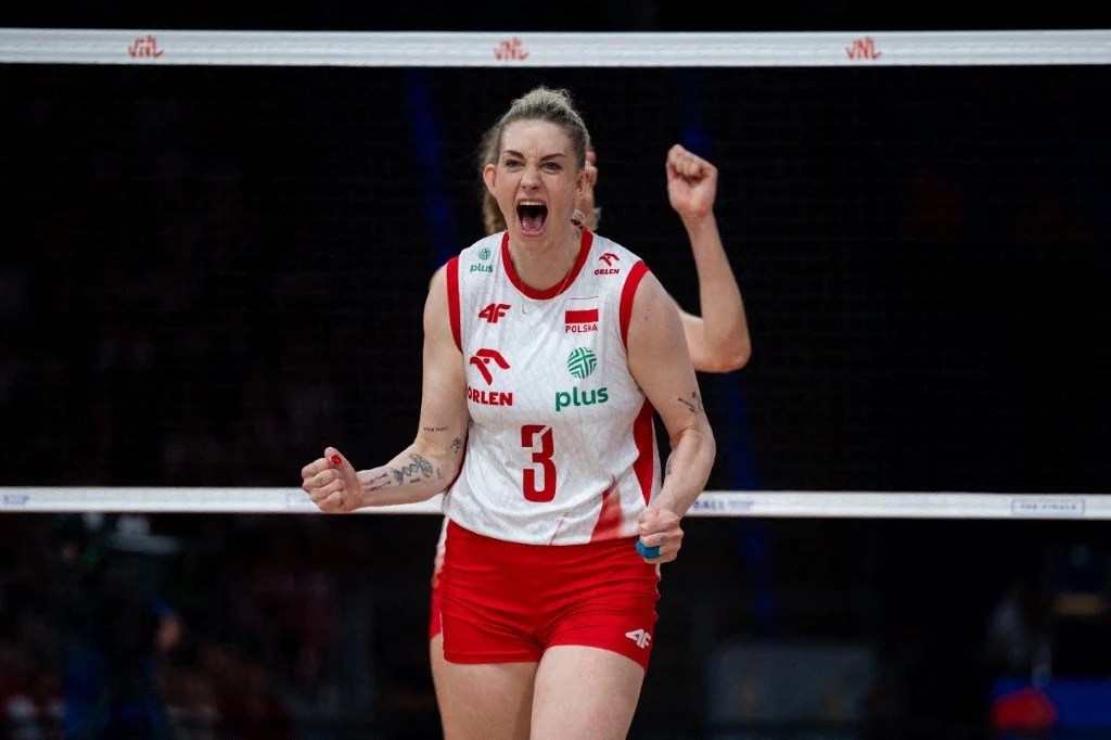 Magdalena Stysiak jest liderką reprezentacji Polski Zawodniczka siatkówki ubrana w strój reprezentacji Polski, z widocznym numerem trzy, wyraża emocje po zdobytym punkcie, trzymając zaciśnięte pięści i otwarte usta, za nią widać fragment siatki oraz rękę drugiej zawodniczki w tle.