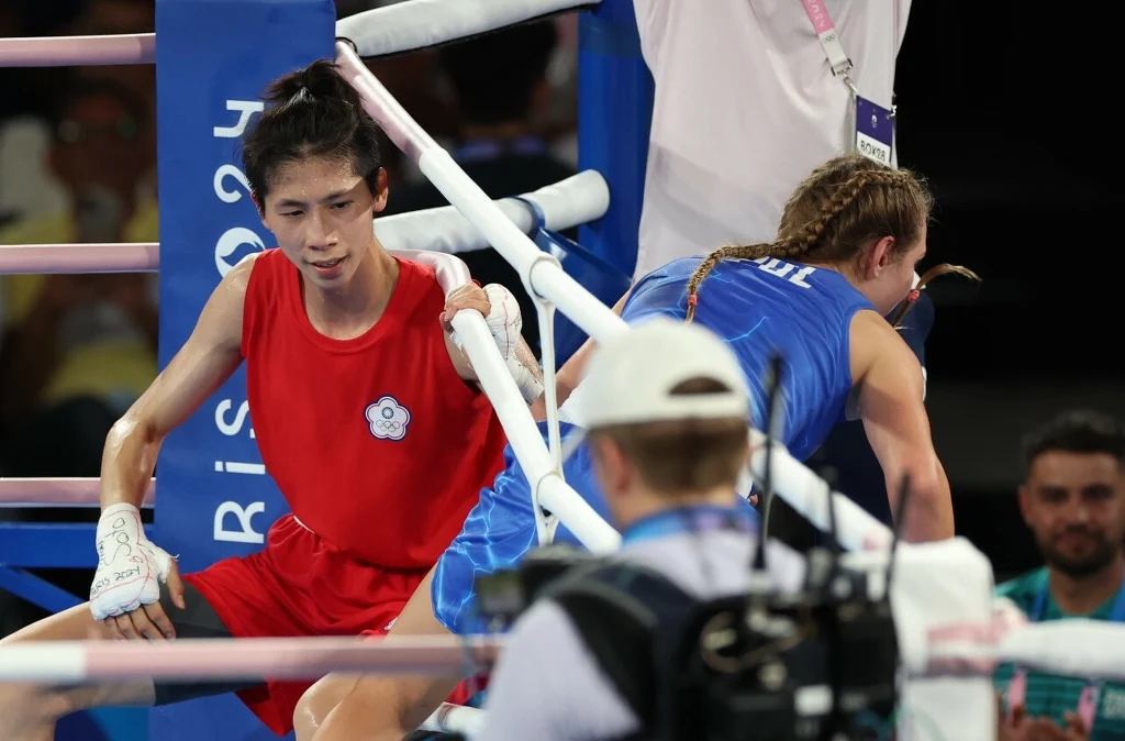 Lin Yu-ting i Julia Szeremeta w finale IO 2024 Dwóch bokserów w ringu, jeden w czerwonym stroju oparty o liny, drugi w niebieskim schylony, zbliżenie na emocje i wysiłek podczas sportowej rywalizacji, w tle sędzia i obserwatorzy.