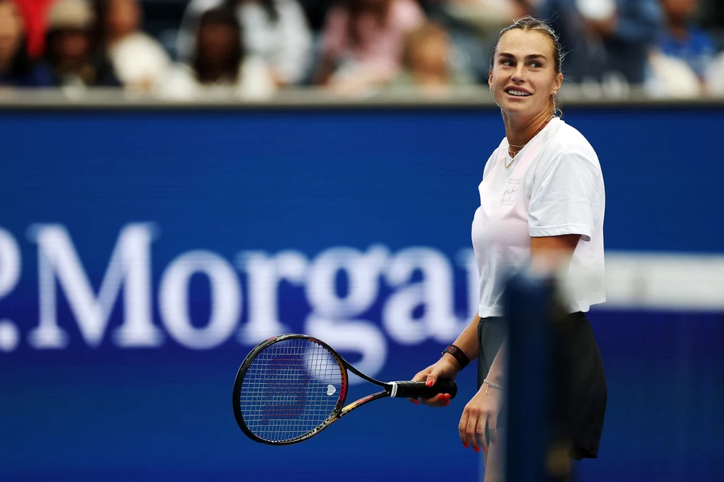Aryna Sabalenka Tenisistka na korcie trzymająca rakietę tenisową, uśmiecha się i patrzy w bok, w tle widownia oraz reklama JP Morgan.
