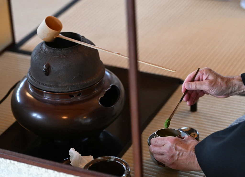Tradycyjne przyrządzanie matchy w Japonii Ceremonia parzenia herbaty z tradycyjnym żelaznym czajnikiem, bambusową łyżką i dwiema rękami przygotowującymi zieloną herbatę, wszystko na tle maty tatami.