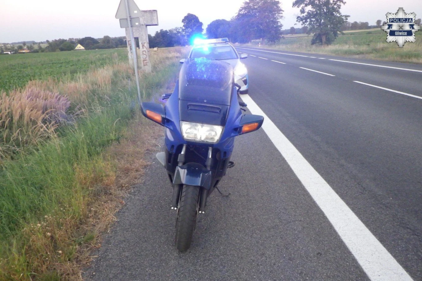 Ponad 2 promile alkoholu w organizmie i 400 km do przejechania. Policjanci zatrzymali pijanego motocyklistę. Motocykl zatrzymany na poboczu przez radiowóz policyjny z włączonymi niebieskimi światłami na wiejskiej drodze, otoczony polami i drzewami.