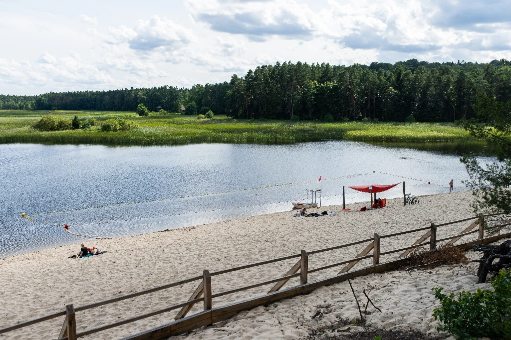 Piaszczysta plaża nad jeziorem, kilka osób odpoczywających przy brzegu, zacienione miejsce z czerwonym zadaszeniem, spokojna woda otoczona zielonym lasem i roślinnością, drewniany płot w pierwszym planie.