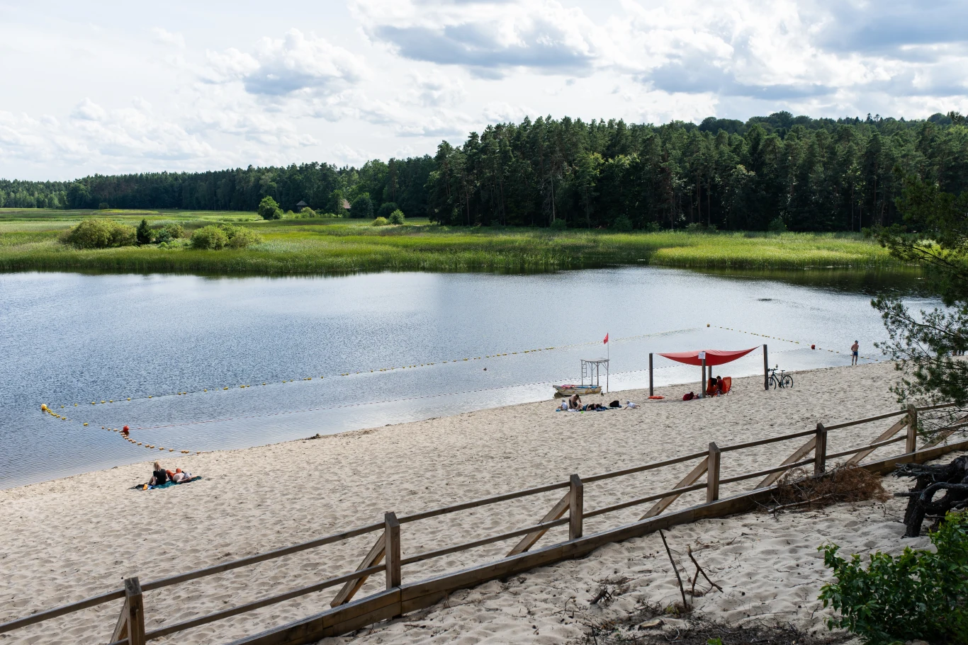 Piaszczysta plaża nad jeziorem, kilka osób odpoczywających przy brzegu, zacienione miejsce z czerwonym zadaszeniem, spokojna woda otoczona zielonym lasem i roślinnością, drewniany płot w pierwszym planie.