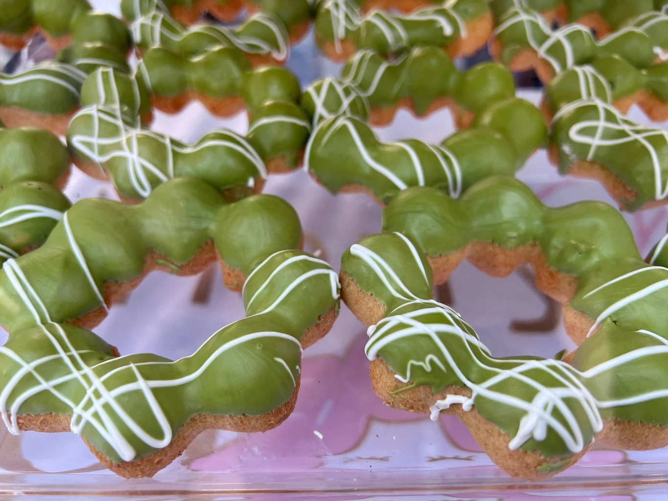 Rząd zielonych donutów polanych polewą matcha z białymi dekoracjami z lukru, ułożonych na przezroczystej tacy