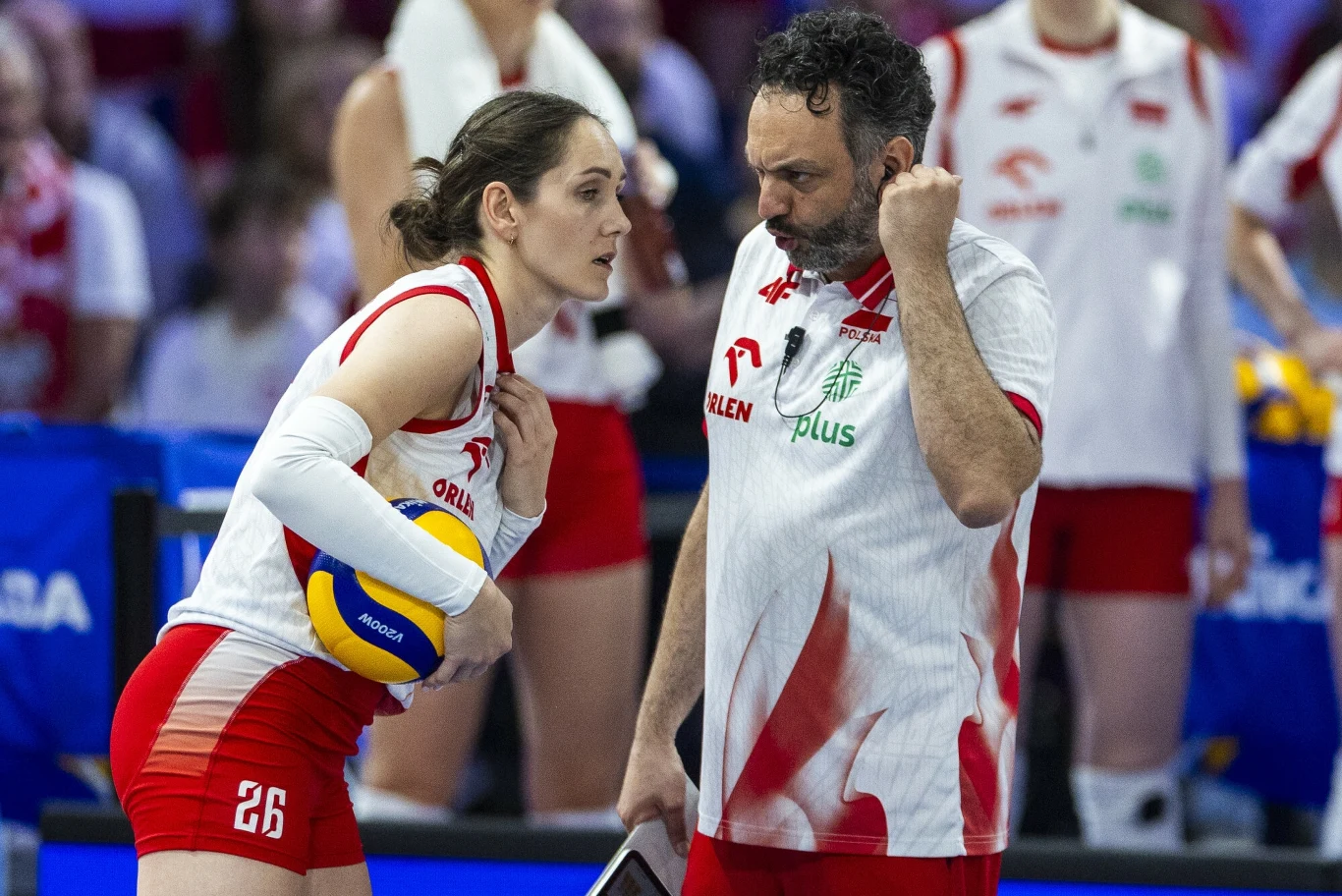 Siatkarka w stroju meczowym trzyma piłkę i rozmawia z trenerem drużyny narodowej, oboje skupieni na strategii gry; w tle widoczne osoby związane z zespołem.
