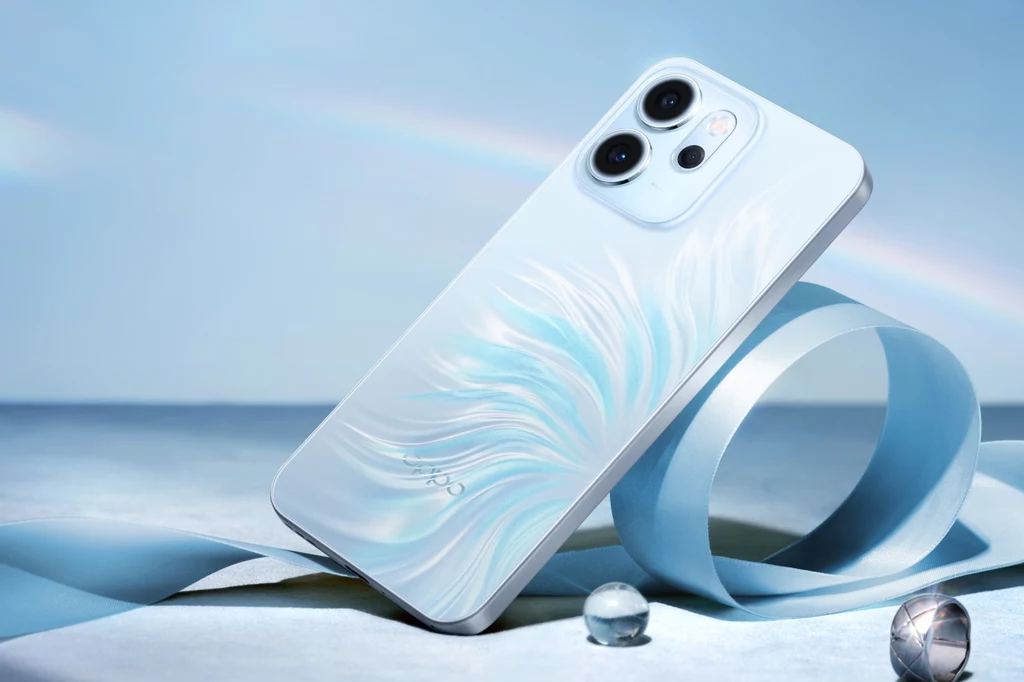 Smartfon Oppo Reno 14 FS 5G. Nowoczesny smartfon o białej obudowie ze wzorem przypominającym delikatne, błękitne pióra, ustawiony na jasnym tle z niebieską taśmą i szklanymi kulkami, w tle widoczna subtelna tęcza.
