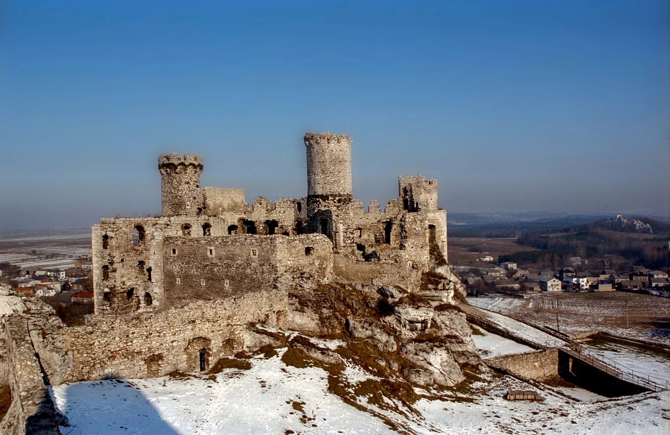 Ruiny średniowiecznego zamku wznoszące się na wzgórzu otoczonym częściowo śniegiem, z panoramicznym widokiem na okoliczne pola i niewielką miejscowość w tle.