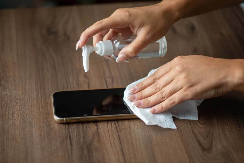 Ręce czyszczące smartfon za pomocą płynu dezynfekującego i chusteczki na drewnianym stole.