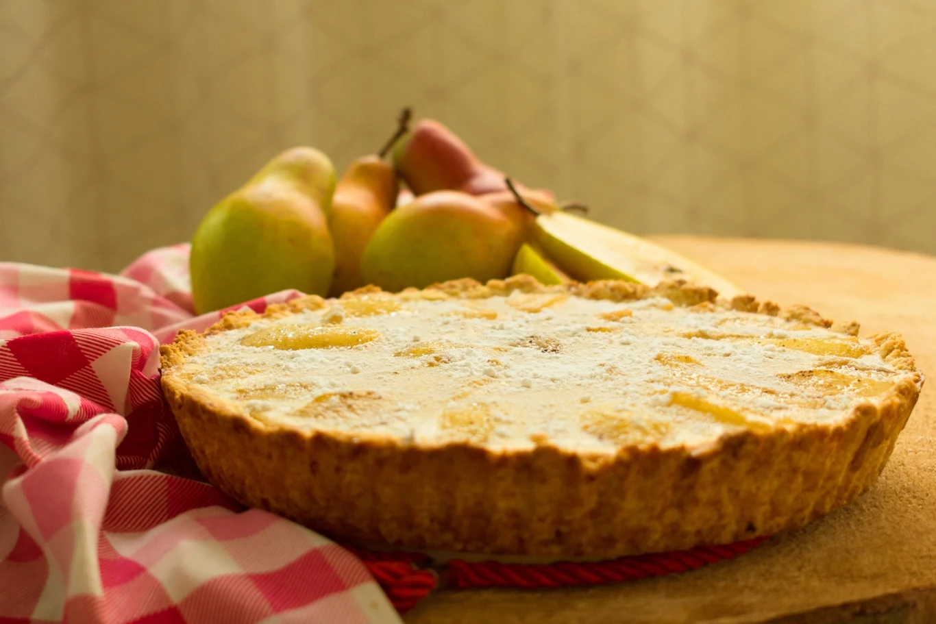 Tarta z jasną polewą posypaną cukrem pudrem ustawiona na stole obok czerwono-białej ściereczki, w tle kilka dojrzałych gruszek.