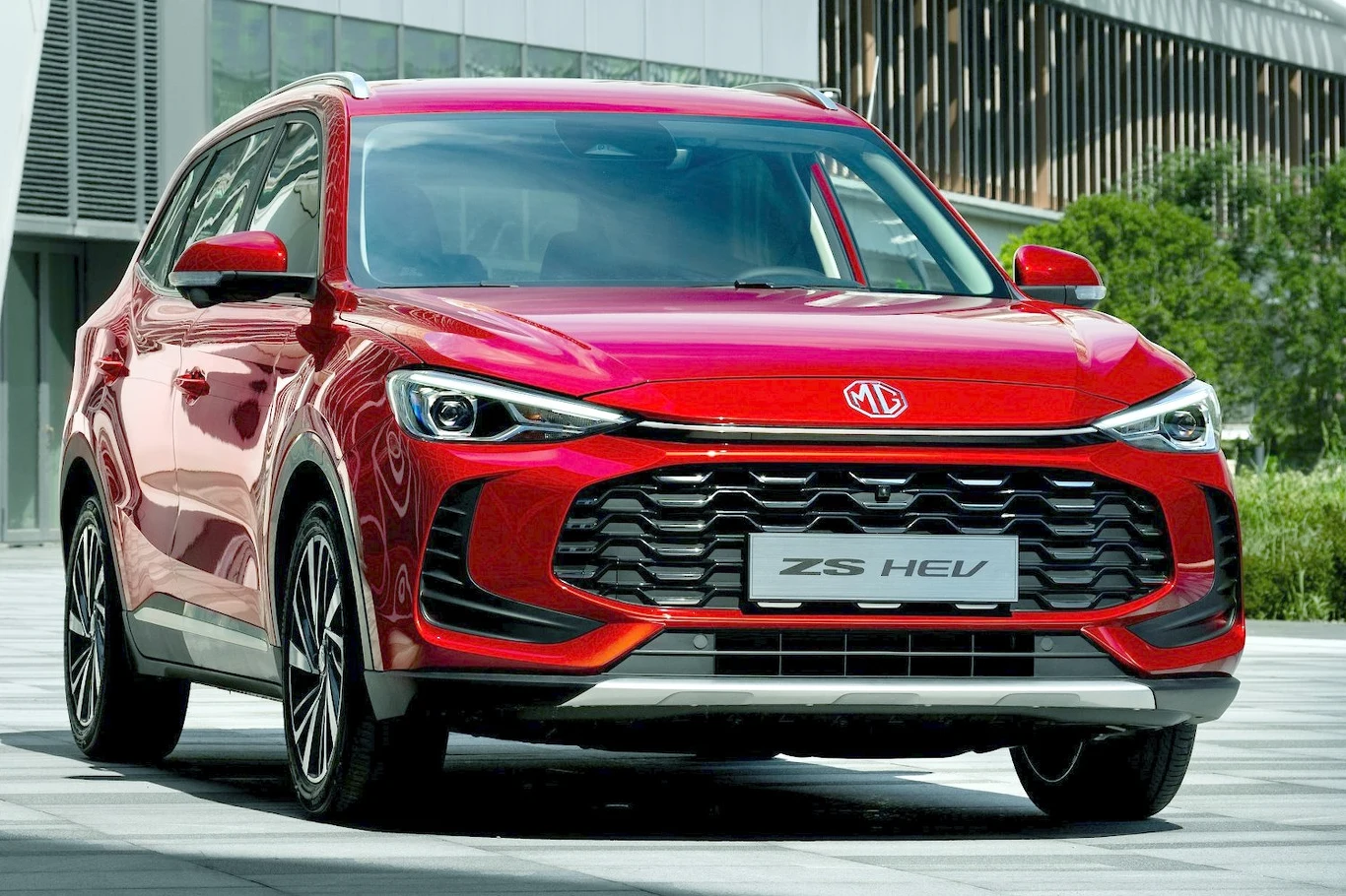 Czerwony nowoczesny SUV MG ZS HEV z charakterystycznym grillem i dynamiczną linią nadwozia, zaparkowany na tle miejskiej architektury i zieleni.