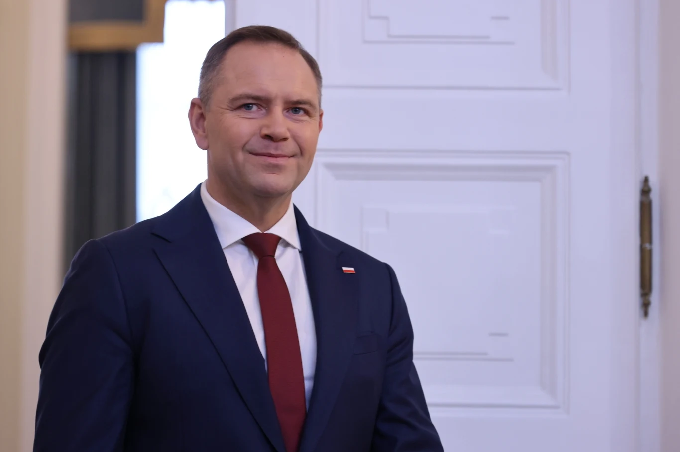 Prezydent Polski Karol Nawrocki Mężczyzna w granatowym garniturze, białej koszuli i bordowym krawacie stoi przed jasnymi drzwiami, przypięta flaga Polski do klapy marynarki.