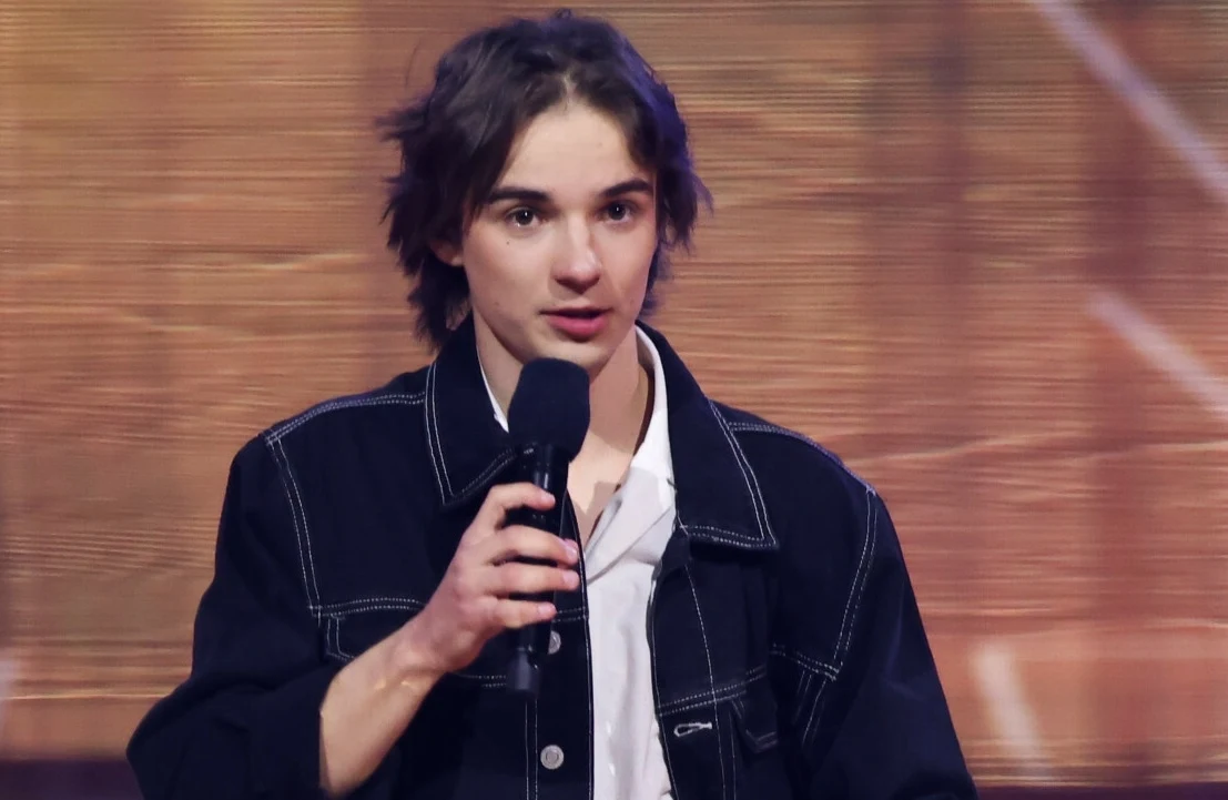 Wiktor Waligóra Młody mężczyzna z średniej długości włosami ujęty podczas przemawiania do mikrofonu, ubrany w ciemną kurtkę z jasną koszulą, w tle rozmyte, ciepłe światło sceniczne.