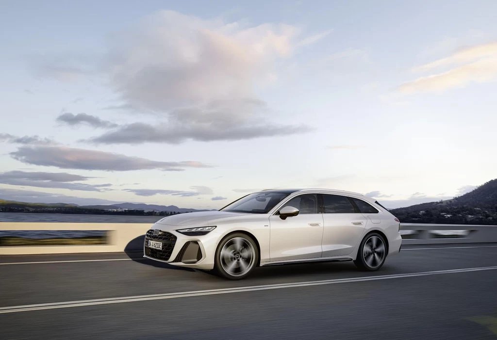 Zużycie paliwa w cyklu mieszanym A6 Avant e-hybrid nie przekracza 3 l/100 km Srebrny samochód osobowy typu kombi poruszający się po drodze na tle krajobrazu z wodą i górami w oddali, przy zachmurzonym niebie.
