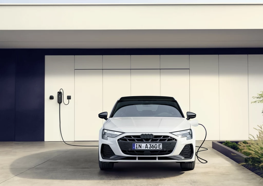 Przyspieszenie od 0 do 100 km/h? W przypadku A3 Sportback TFSI e to zaledwie 6,3 sekundy Biały samochód marki Audi podłączony do ładowarki elektrycznej przy nowoczesnym garażu, widok z przodu, betonowy podjazd, minimalistyczna architektura.