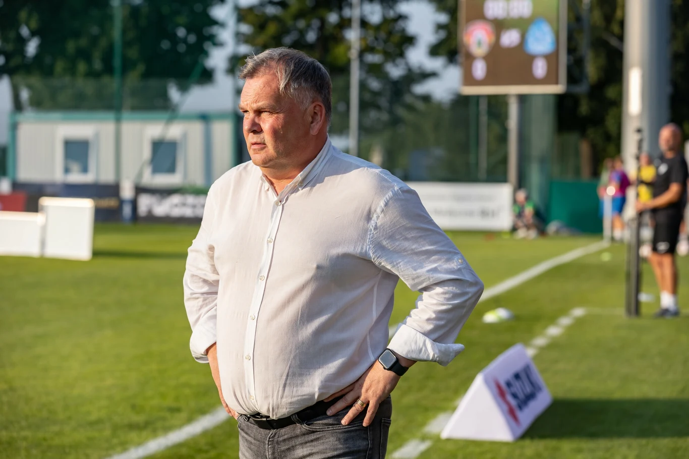 Mężczyzna w białej koszuli stoi przy linii bocznej boiska piłkarskiego, obserwując wydarzenia na murawie, w tle widoczne są tablice wyników i kilka osób.