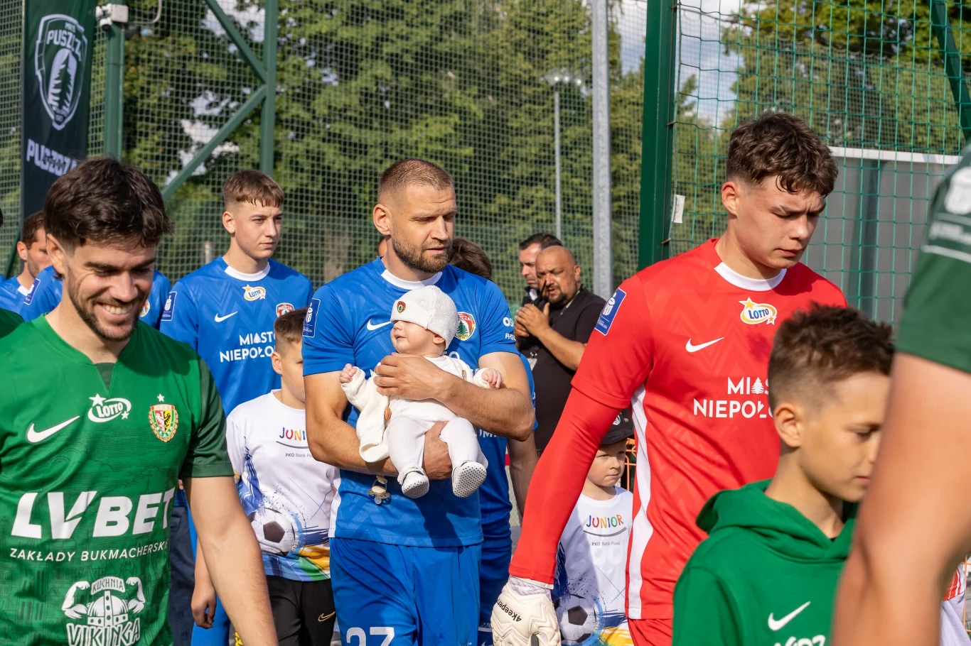 Piłkarze wychodzący na murawę podczas wydarzenia sportowego, na pierwszym planie zawodnik w niebieskim stroju trzyma na rękach niemowlę, obok inni sportowcy w zielonych i czerwonych koszulkach oraz dzieci.
