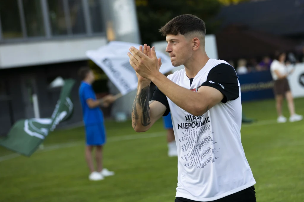 Puszcza Niepołomice, prezentacja strojów Młody mężczyzna w sportowej koszulce z napisem 'NIEPOMALNI' stoi na boisku, z dłońmi złożonymi do oklasków, w tle osoby rozmawiające oraz banery i fragmenty budynku.