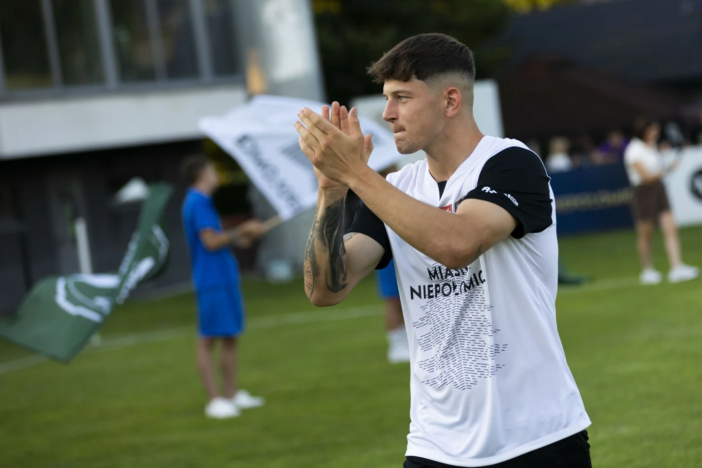 Młody mężczyzna w sportowej koszulce z napisem 'NIEPOMALNI' stoi na boisku, z dłońmi złożonymi do oklasków, w tle osoby rozmawiające oraz banery i fragmenty budynku.