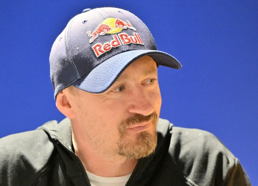 Mężczyzna z krótką brodą i w baseballowej czapce Red Bull na głowie, ubrany w czarną bluzę, patrzący w bok na niebieskim tle.