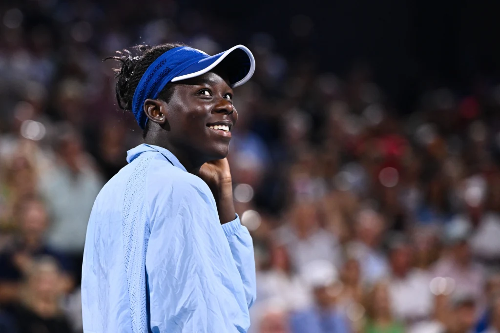 Victoria Mboko Uśmiechnięta młoda kobieta w sportowej odzieży i niebieskiej czapce z daszkiem stoi na tle rozmytego tłumu kibiców podczas wydarzenia sportowego.