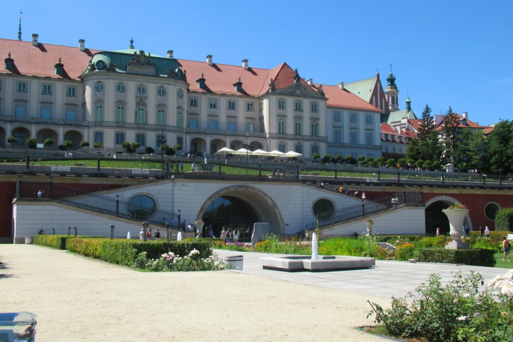 Pałac z ozdobnym dachem i licznymi oknami góruje nad zadbanym ogrodem. Na pierwszym planie widać fontannę otoczoną alejkami z zielenią i kwiatami. W tle pod pałacem duże, łukowate przejście podziemne, a na tarasie pawilony z parasolami.