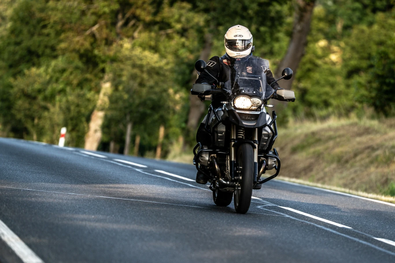 Czy ktoś strzela do motocyklistów? Motocyklista jadący po asfaltowej, pustej drodze w otoczeniu zieleni, ujęcie frontalne, kierowca w kasku i profesjonalnym ubraniu motocyklowym.